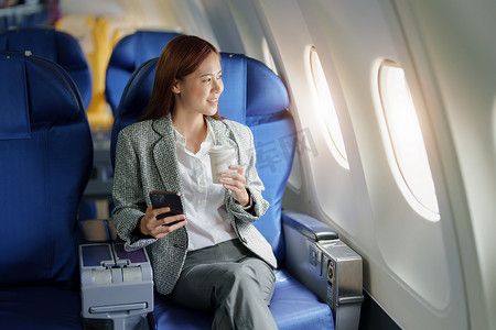 一位成功的亚洲女商人或穿着正式西装的女企业家在飞机上的肖像坐在商务舱座位上，在飞行期间使用智能手机喝咖啡，放松概念