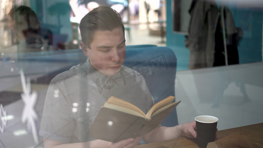 一个年轻人坐在咖啡馆的玻璃杯后面看书。
