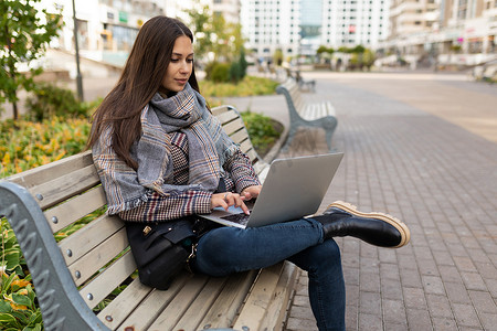 在城市公园的背景下，年轻成功的女性自由职业者穿着温暖的秋装，在笔记本电脑上工作