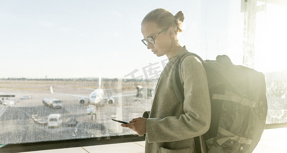 休息大堂摄影照片_年轻女子站在机场登机口窗边，手里拿着手机，背着旅行背包，走到休息区。