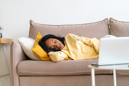 美国客厅摄影照片_无聊的非洲裔美国女性睡在客厅的沙发上，同时在笔记本电脑上播放电影