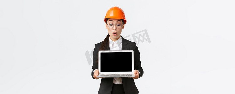 令人印象深刻的亚洲女性建筑工程师、建筑师或企业经理展示笔记本电脑屏幕，表情惊讶，在会议期间查看利润图，在工厂戴安全帽
