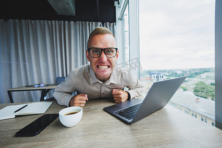 情绪化的男人在工作场所看着相机，一个戴眼镜的男人，坐在电脑前，一个学生正在办公室咖啡馆里寻找新想法的灵感