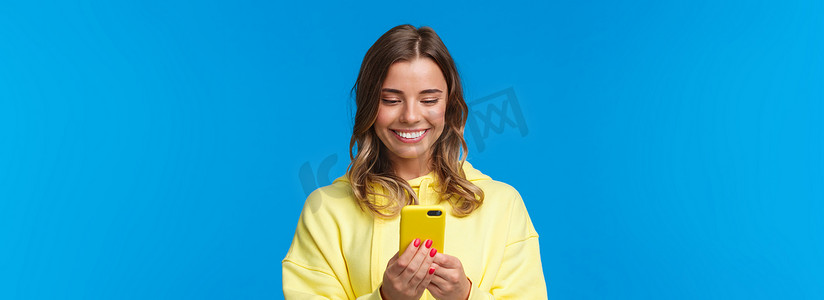 穿着黄色连帽衫的愚蠢美丽金发欧洲女人的特写肖像，微笑着看着手机显示屏，观看可爱的视频，与男朋友发短信，站在蓝色背景