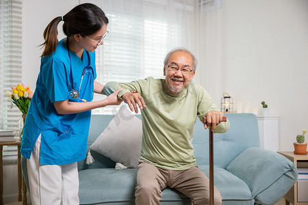 拐杖摄影照片_年轻的护理人员支持帮助老人用沙发上的拐杖站起来