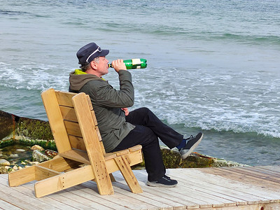 一个男人坐在海边的木制躺椅上喝啤酒