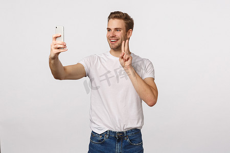 快乐的男人下载新应用程序，尝试可爱的过滤器，在智能手机上自拍，做和平标志，快乐地微笑，给自己拍照，站在白色背景