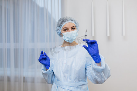 手术帽摄影照片_年轻女医生护士的肖像，身穿白大褂，戴着狭缝面罩手术帽，戴着蓝色医用手套，拿着注射器，看着镜头