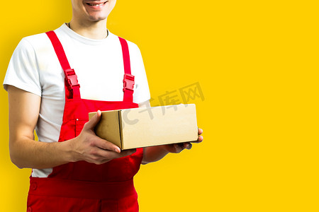 一位戴着红帽的快乐年轻送货员的法师站在黄色背景中隔离的包裹邮箱里