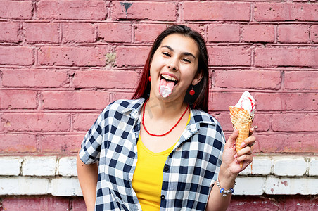 吃冰淇淋的女人摄影照片_在街上粉色砖墙背景上吃冰淇淋的红头发开朗时髦女人