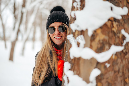 戴着太阳镜和帽子的年轻女子躲在雪地公园的树后