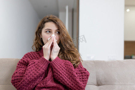 一位年轻女子在家生病，流鼻涕，用纸手帕擦鼻子，坐在沙发上