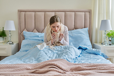 家里生病的年轻女人躺在床上，手里拿着一杯热饮和手帕