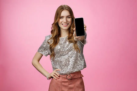 摄影棚拍摄了自信、魅力四射的年轻魅力女性，介绍了很棒的智能手机应用程序，展示了手机显示微笑自信推荐关注博客页面，站在粉红色背景