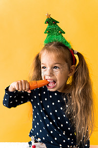 小鹿女孩摄影照片_肖像正面开朗微笑快乐的小女生女孩圣诞树装饰波尔卡圆点连衣裙咬吃橙色背景上的橙色胡萝卜。