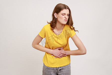 身穿休闲T恤、棕色头发的不快乐患病少女站着，用手托着肚子、胃痉挛或经期疼痛的肖像。