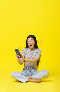 兴奋的亚洲年轻女孩坐在地板上，拿着智能手机发短信或网上购物，或赢得黄色背景下的游戏。