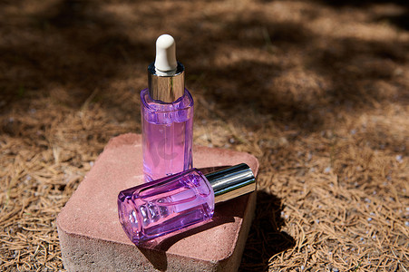 化妆品小滴管摄影照片_石头上有抗衰老血清的紫罗兰色小瓶，在落下的干松树针上，有阳光和针叶树阴影