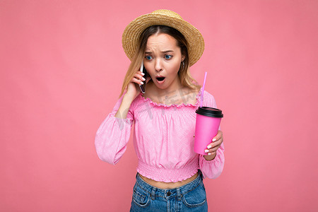 照片中，美丽的、震惊的、情绪激动的年轻金发女子穿着粉色短上衣、头戴草帽，一边喝着粉红色背景的饮料，一边打电话，还有复制空间