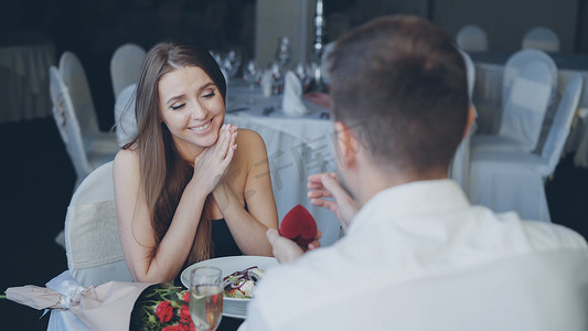 帅哥向惊讶的漂亮女人求婚，然后在餐厅浪漫约会时将订婚戒指戴在她的手指上并亲吻她的手。