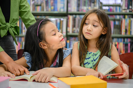 小孩儿看书摄影照片_两个可爱的女孩在老师教学时在图书馆看书时互相嫉妒。