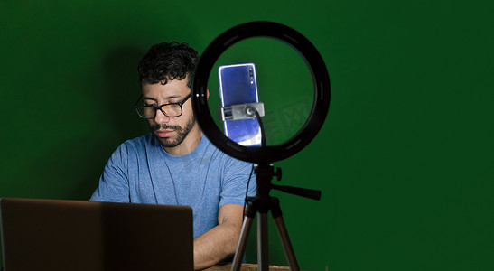 手持铃摄影照片_用笔记本电脑制作视频博客的年轻人，用手机制作内容的年轻人创作者，用笔记本电脑进行绿色背景视频通话的男人，