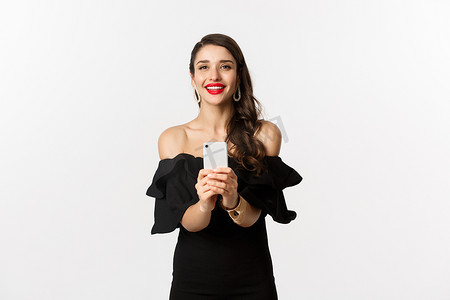 迷人的派对女孩在智能手机上拍照，在手机相机上拍照，穿着黑色裙子站在白色背景上
