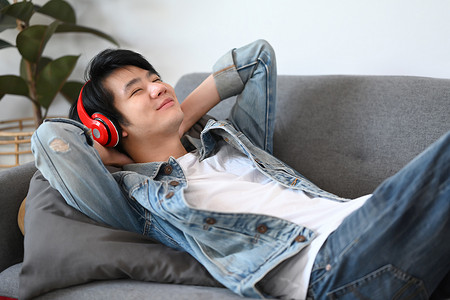 快乐的亚洲男人用无线耳机听音乐，躺在家里的沙发上。