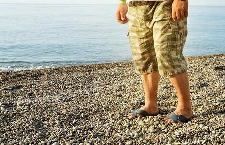 等待日出摄影照片_男人的脚穿着人字拖和短裤，一个男人站在卵石滩的海滩上，早晨的日出