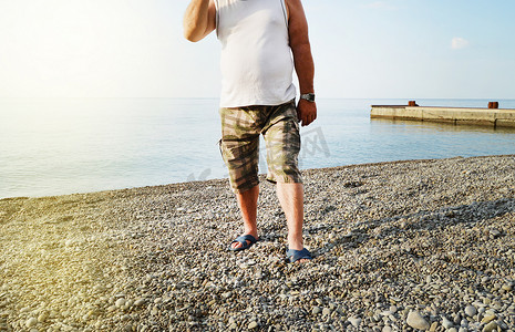 男人的脚穿着人字拖和短裤，一个男人站在卵石滩的海滩上，早晨的日出