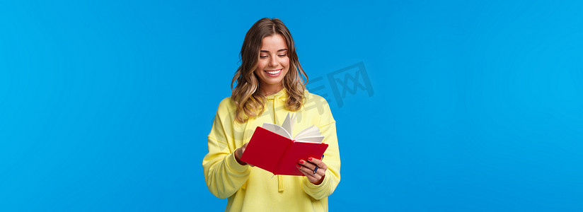女孩买了新的计划书，写下笔记或演讲，开心地笑着，拿着红色笔记本，决定开始秘密日记，站在蓝色背景中的黄色连帽衫