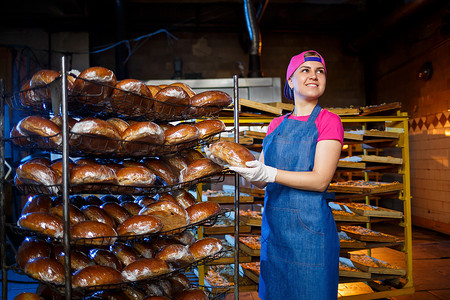 拿着面包的女孩摄影照片_专业面包师 — 一位穿着牛仔裤围裙的年轻漂亮女人在面包店或面包店的背景下拿着新鲜的面包。