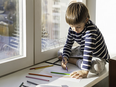 彩虹蜡笔手绘摄影照片_小男孩坐在窗台上，用彩色铅笔画彩虹。