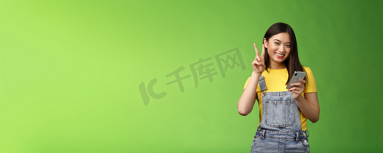 可爱温柔的亚洲女性拿着智能手机，展示胜利的和平标志，看起来很高兴，在绿色背景附近摆出无忧无虑的姿势，赢得在线赠品，在比分游戏中获胜