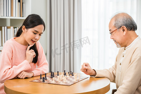 美丽的年轻微笑女人坐在家里与年长的老人下棋，玩得很开心