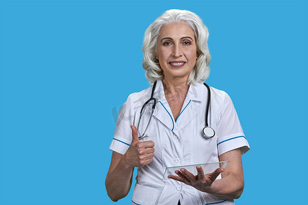 微笑的高级医生使用透明数字平板电脑，并在蓝色背景上显示拇指手势。