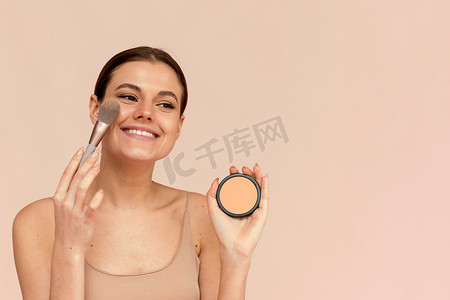 一个美丽的女人在粉红色的背景下用化妆刷在她的脸上涂抹粉末。