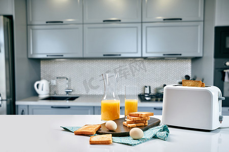 浅色厨房桌上的烤面包机，配有新鲜面包、鸡蛋和一杯橙汁