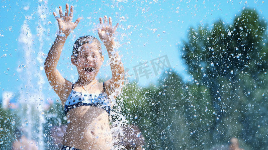 溅水摄影照片_微笑、快乐的八岁女孩穿着泳衣，在街头城市喷泉、户外、公园、夏天、阳光明媚的假期里享受泼水的乐趣。