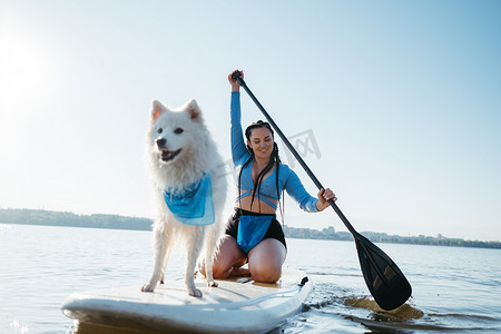 快乐的女人带着她的宠物在城市湖上划桨，雪白的日本斯皮茨狗站在 Sup 板上