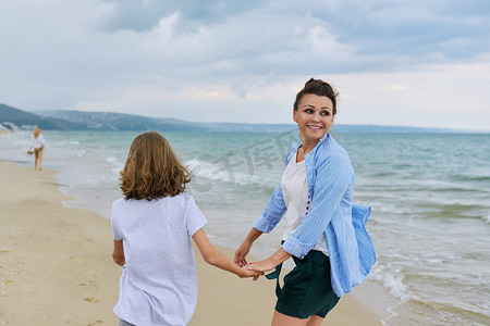 母亲牵孩子背影摄影照片_快乐的妈妈和女儿手牵着手走在沙滩上，背影