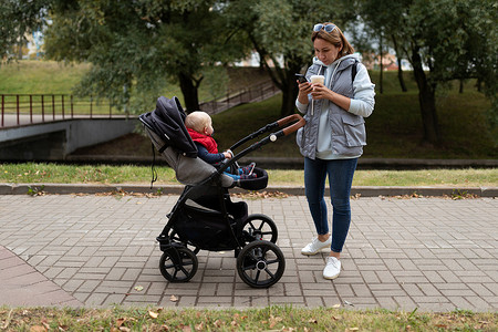 婴儿推车摄影照片_一位年轻的母亲一边喝咖啡一边看手机，带着婴儿推车在城市里散步