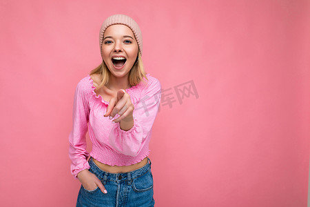 照片中，年轻积极快乐的金发美女带着真挚的情感，身穿粉红色衬衫和粉红色帽子，在粉红色背景中突显出来，有复制空间，手指对着相机