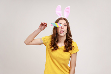 穿着黄色休闲 T 恤、长着粉红色兔耳朵的可爱风趣少年的画像，看着相机，用冰淇淋遮住眼睛。