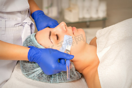 在水疗美容院的皮肤护理过程中，美容师用保湿清洁面膜覆盖女性面部皮肤。