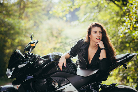 冷淡摄影照片_在阳光明媚的天气里，在大自然、街道、森林里，一位穿着黑色皮衣骑着摩托车的漂亮年轻女子的肖像，特写，看着相机