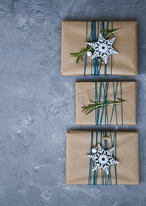 三件用牛皮纸包裹的圣诞礼物，蓝色线包裹着，灰色混凝土背景上有白色雪花和迷迭香枝条。