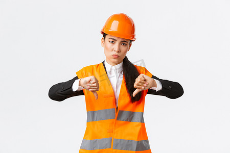 失望而心烦意乱的亚洲女总工程师检查糟糕的工作，表现出失望的大拇指，戴上安全帽，抱怨建筑师的糟糕工作，白色背景