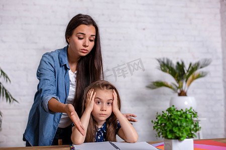 沮丧孩子摄影照片_妈妈坐在家里的桌子旁做作业时对女儿感到沮丧，因为女儿在学习困难的作业。
