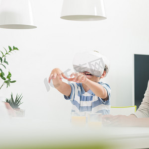 惊讶的孩子坐在餐桌旁，使用虚拟现实耳机试图抓住不存在的虚拟物体。